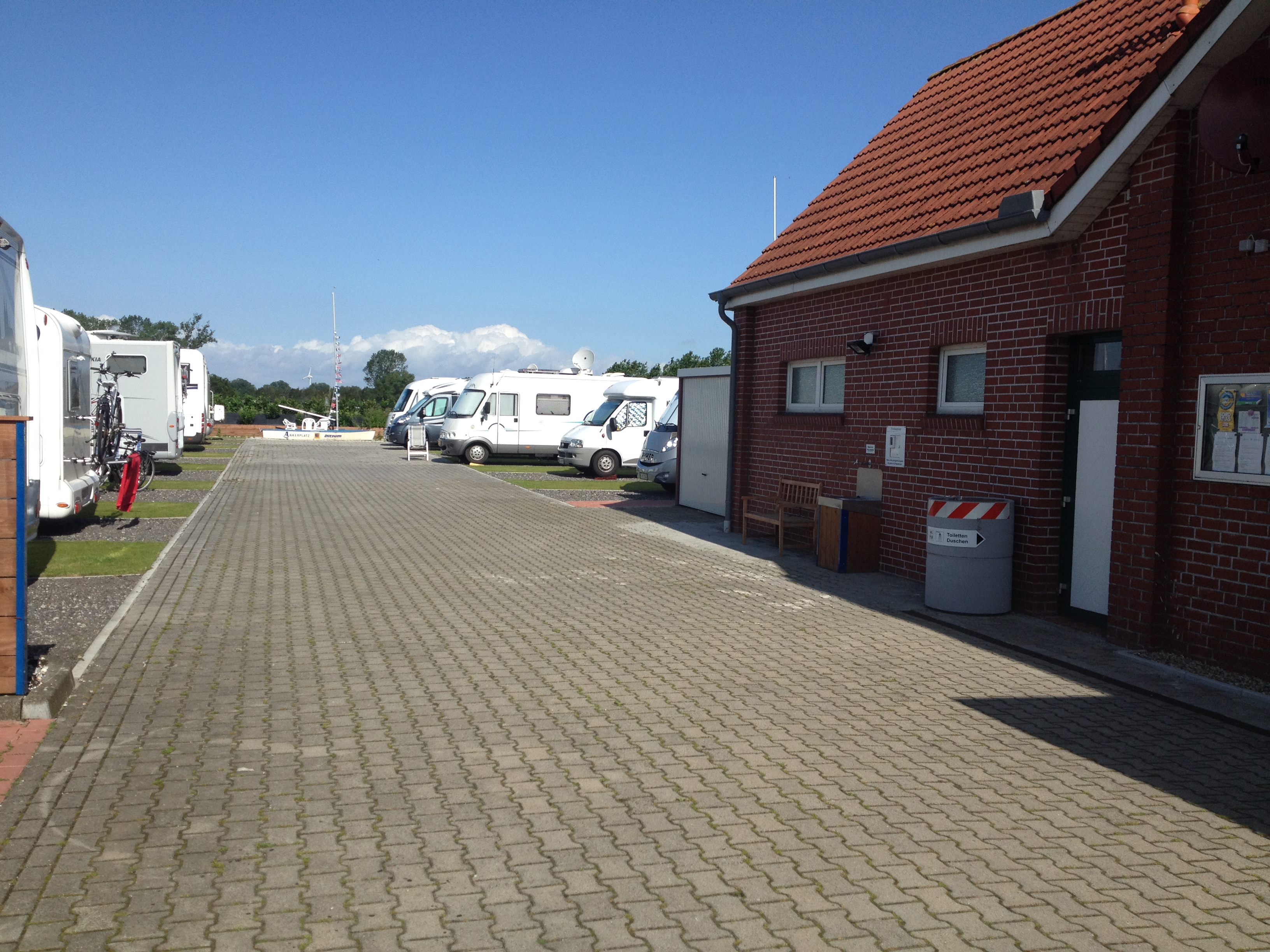 Place de parking pour camping-car - Stromanschluss - Allemagne - Auf der rechten Seite sehen sie hinter der Informationstafel den Eingang zum Sanitärgebäude.
Im Anschluss finden sie die Trinkwasserversorgung mit einem Waschbecken und eine Bank auf der es sich gemütlich sitzen läßt. Die Wlan-Antenne ist deutlich sichtbar. - Ankerplatz Blank Ditzum